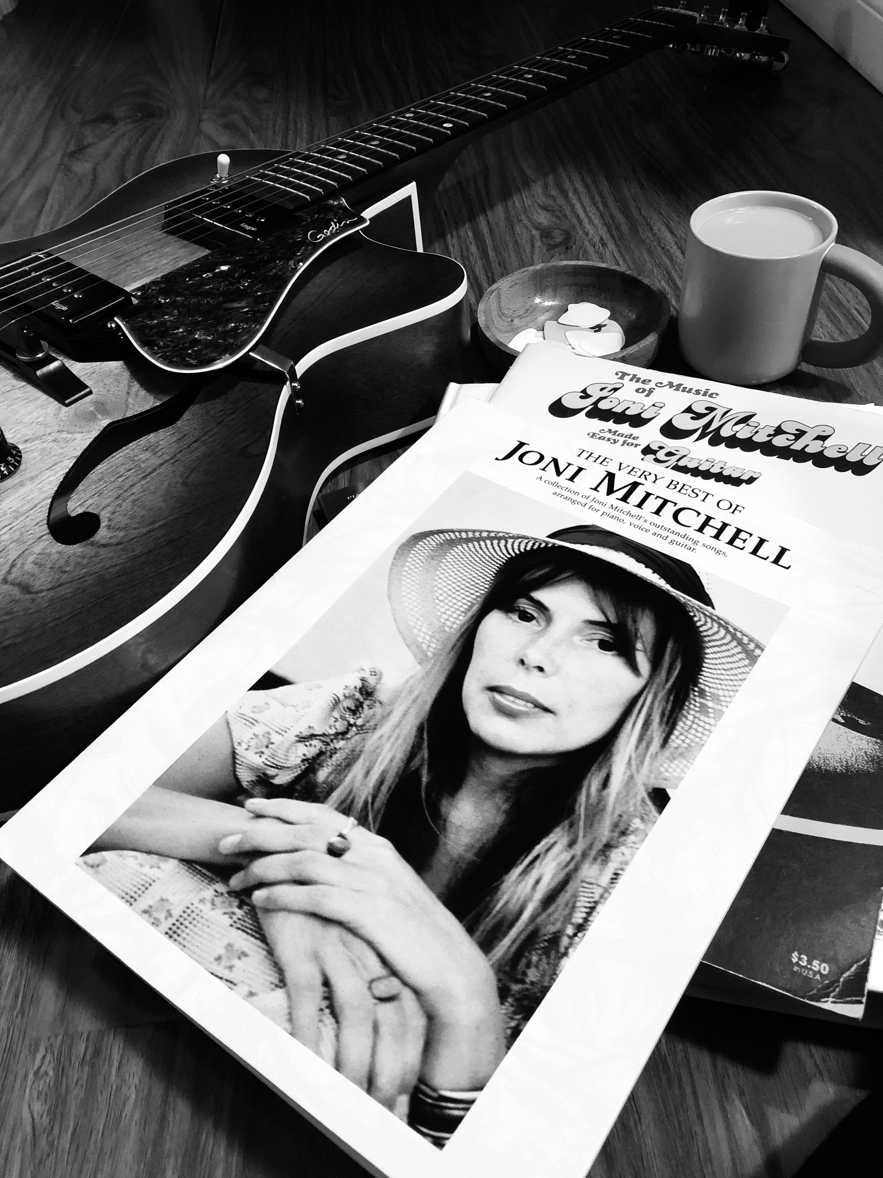 A guitar, Joni Mitchell songbooks and a mug on a tabletop, in black and white